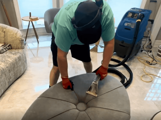 Professional house cleaner expertly steam-cleaning a fabric ottoman, restoring freshness and removing stains using eco-safe methods.